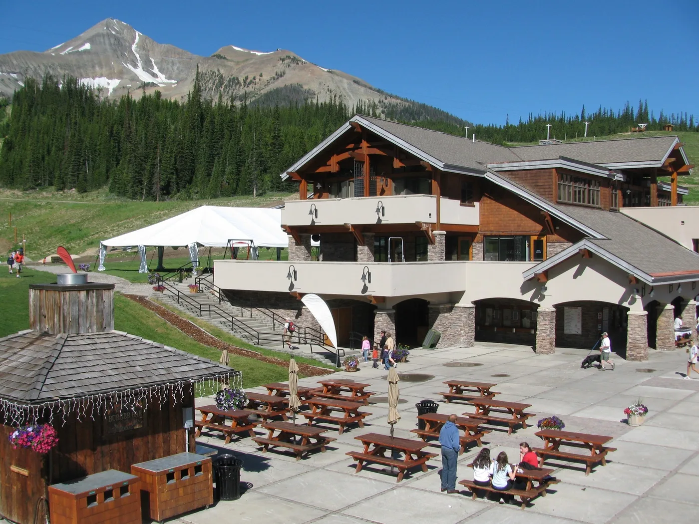 A mountain lodge stands beside a paved patio with picnic tables and a white tent.