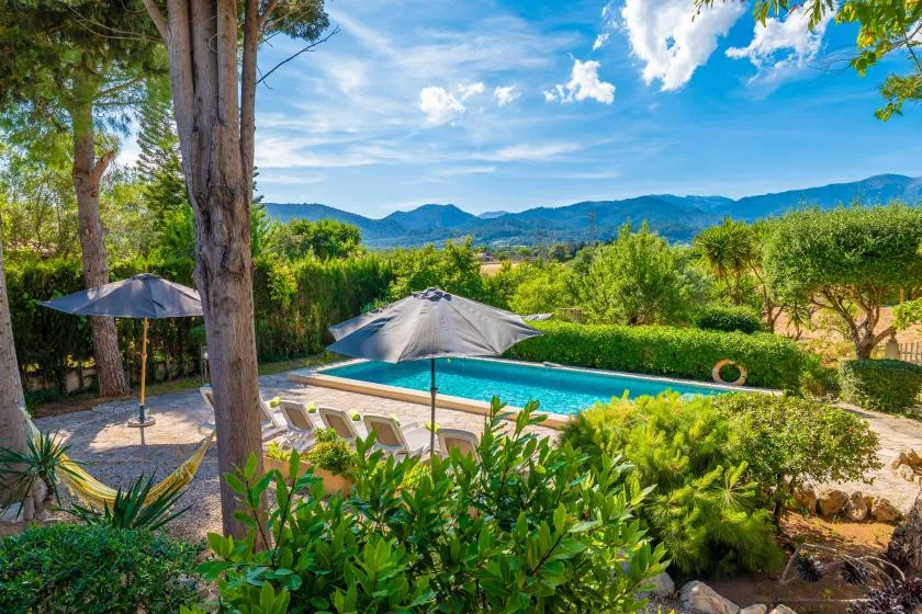 An outdoor swimming pool sits among trees and greenery, with hills in the distance.
