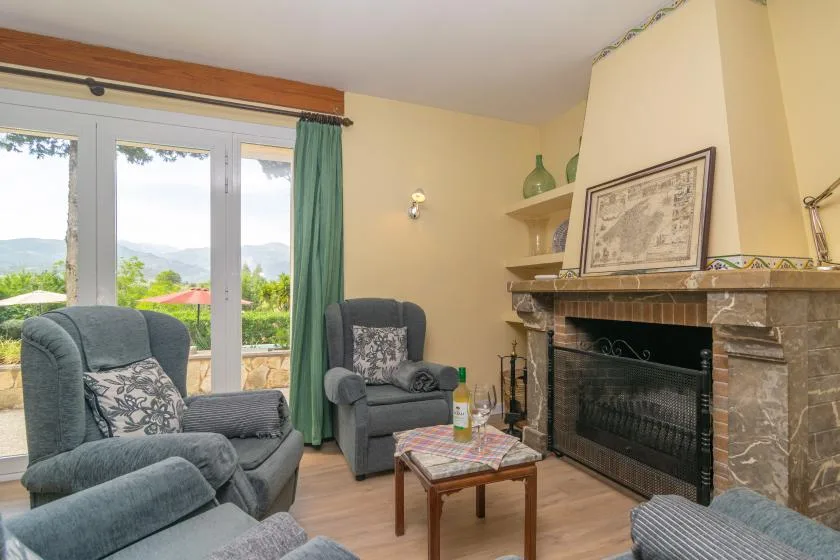 A living room with cushioned seating, a coffee table, and a fireplace beside large windows.