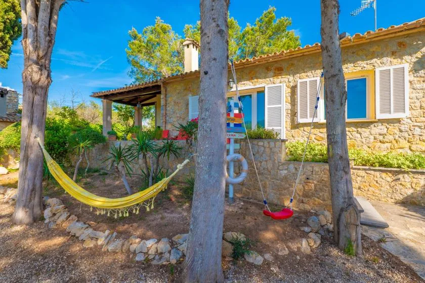 A stone house sits behind a garden with a hammock and swing.