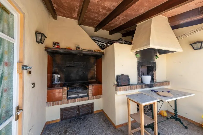 A rustic indoor cooking area with a large fireplace and a central worktable.