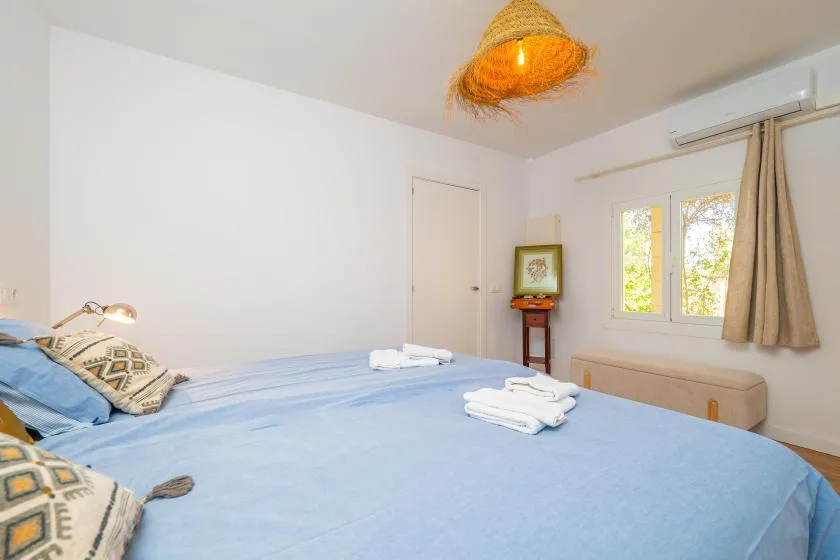 A bright bedroom has a blue bed, a window, and a small bedside table.