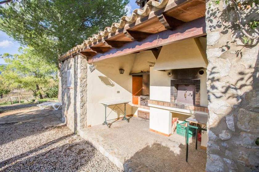 An outdoor cooking area sits beneath a tiled roof beside a stone wall.