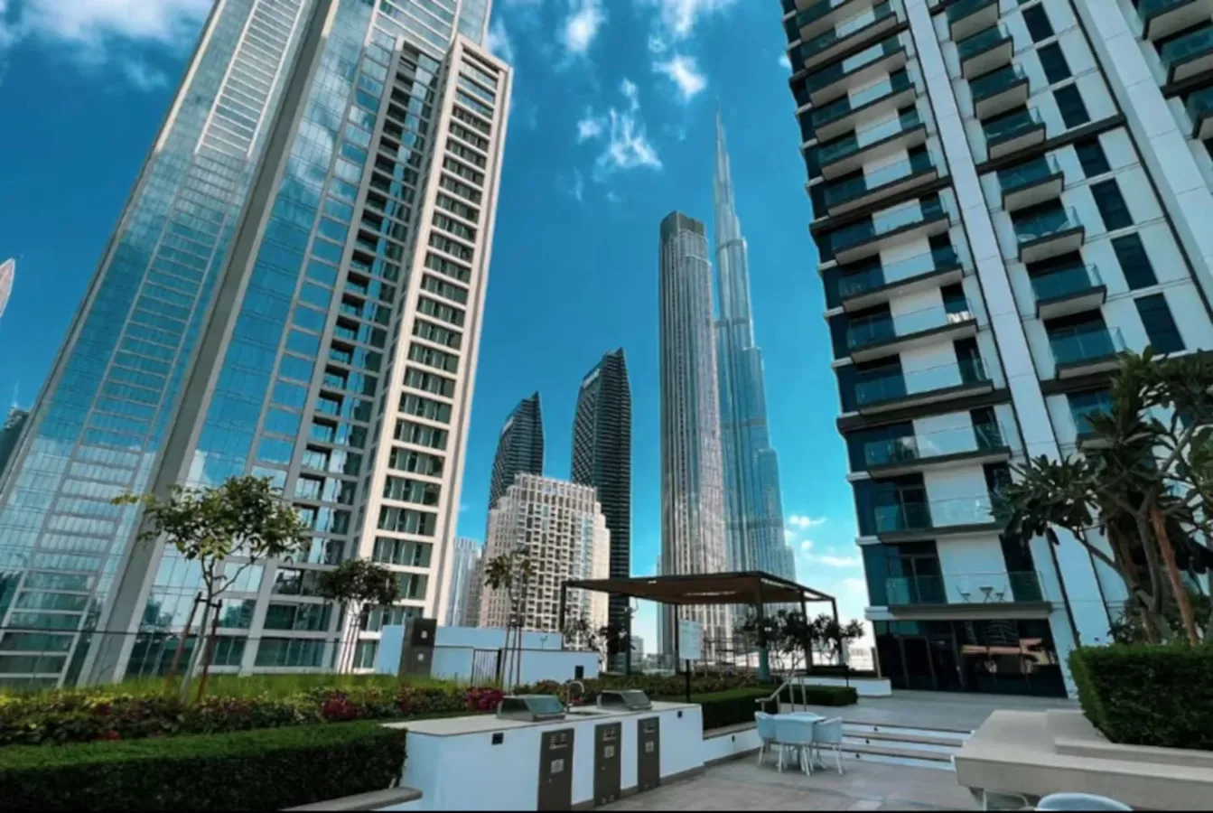 Edificios modernos de gran altura rodean un patio ajardinado al aire libre bajo un cielo azul.