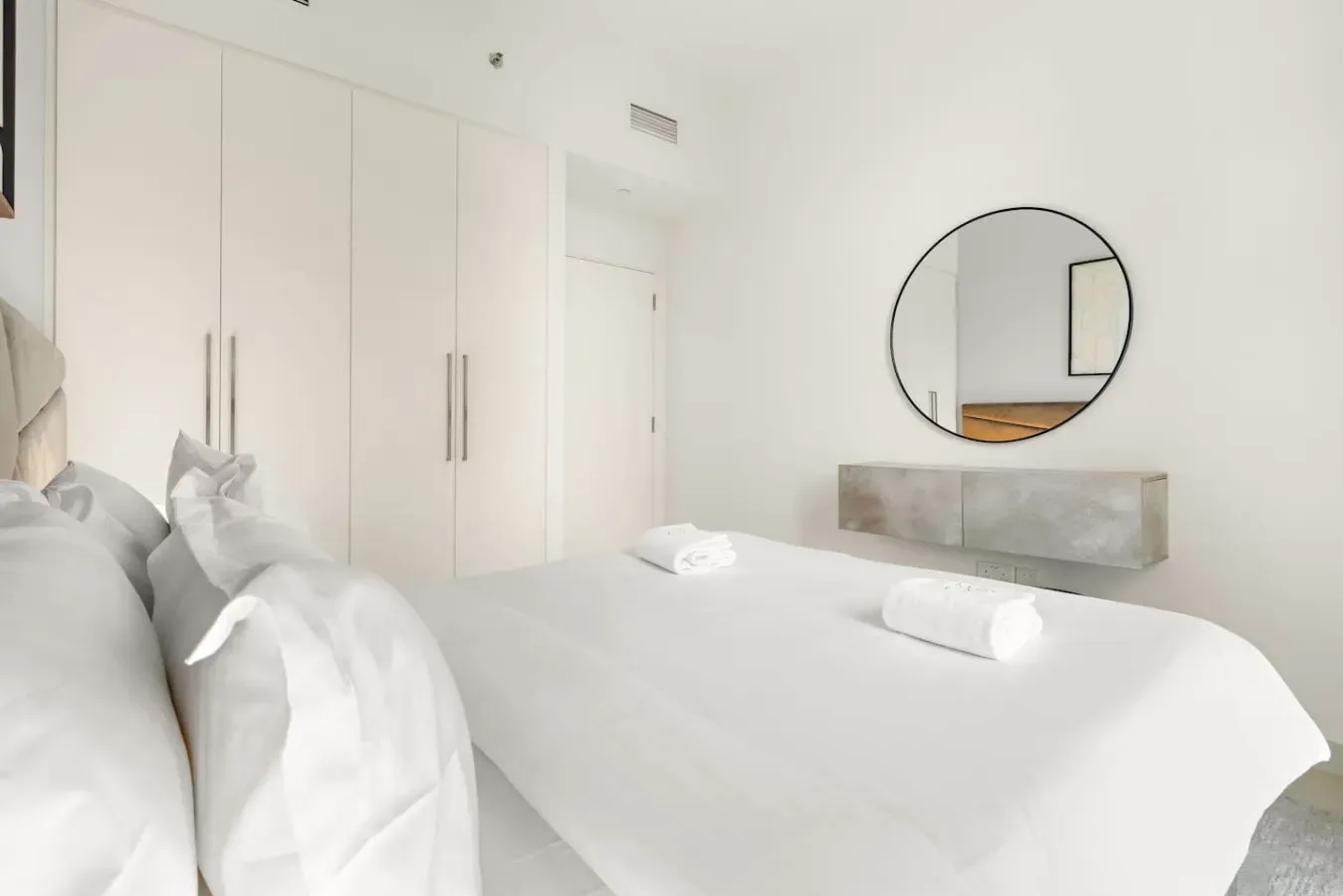 Un dormitorio luminoso con ropa de cama blanca, armarios empotrados y un espejo redondo.