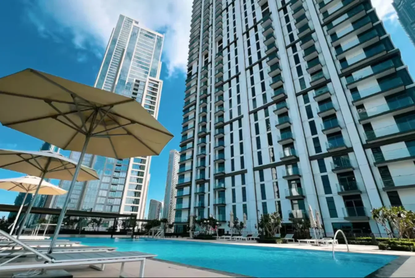 Un edificio alto da a una piscina exterior y una zona de asientos con sombra.