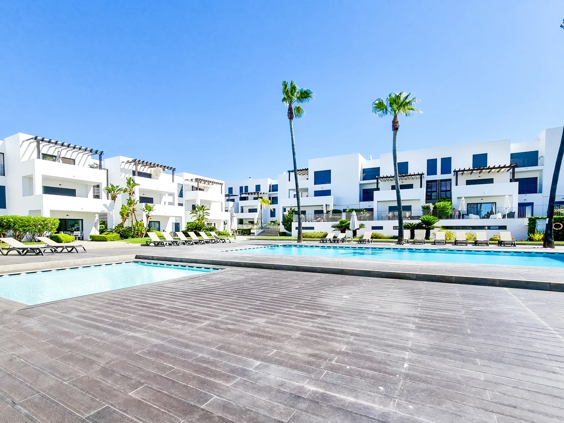 Edificios blancos modernos rodean una piscina exterior y una zona pavimentada junto a ella.