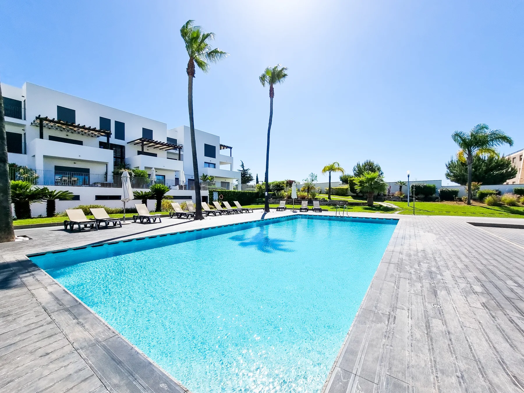 Una piscina al aire libre está junto a edificios modernos y una terraza pavimentada.