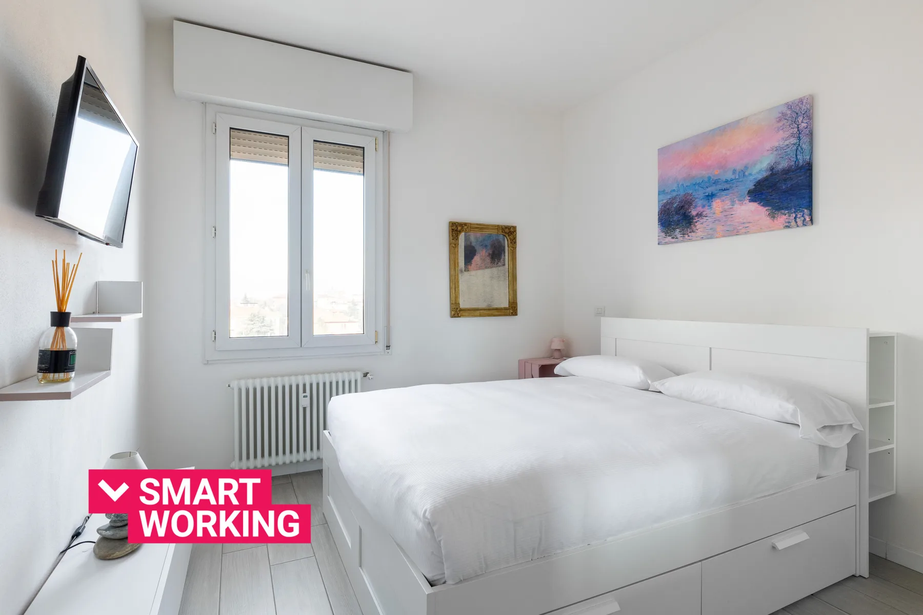 Un dormitorio luminoso con una cama blanca, una ventana y cuadros coloridos.