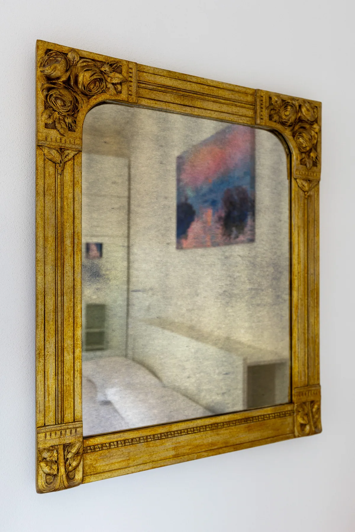 Un espejo con marco ornamentado cuelga en una pared blanca y refleja una habitación con una cama y una obra de arte.