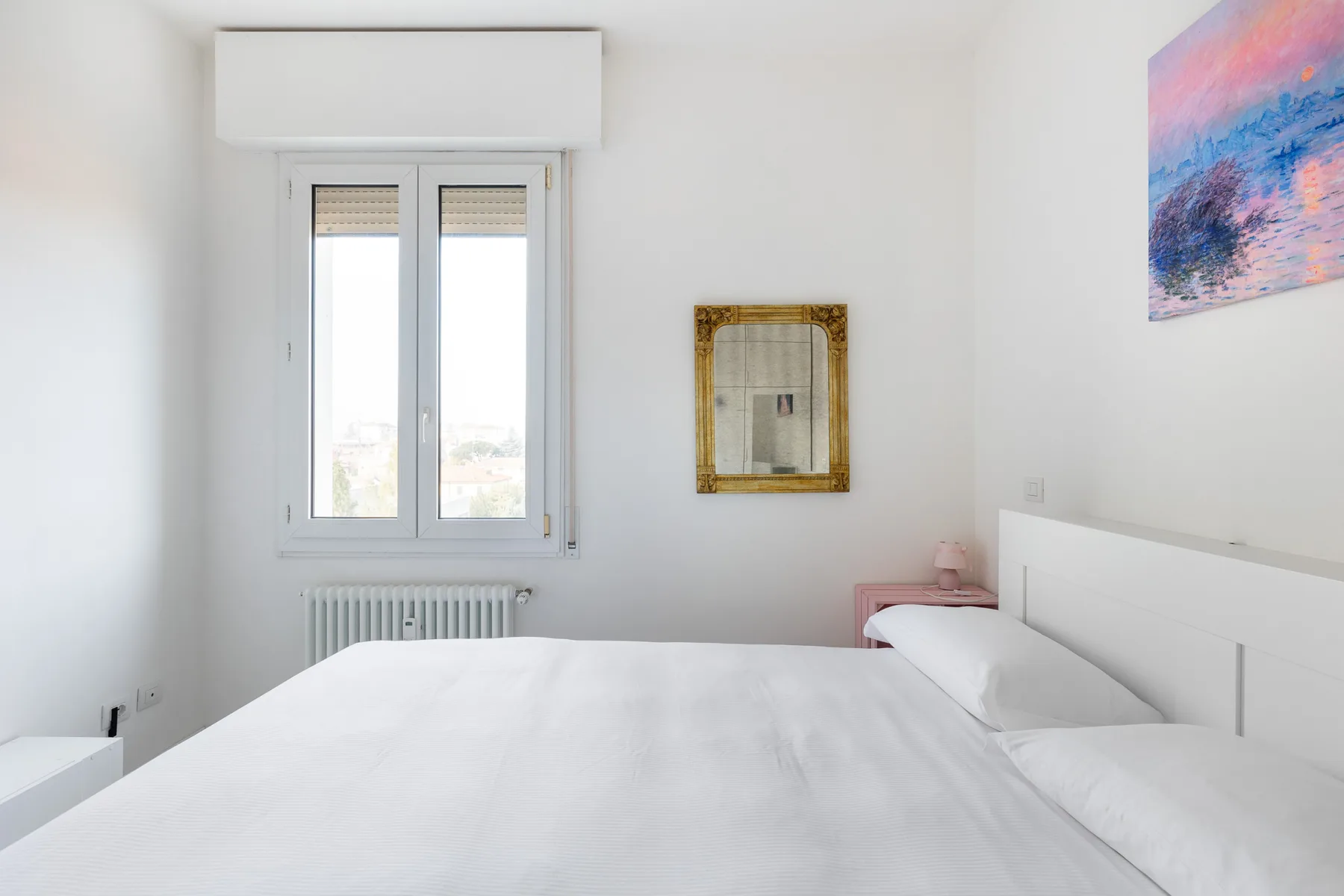 Un dormitorio luminoso con una cama bien tendida, ventanas y arte colorido en la pared.