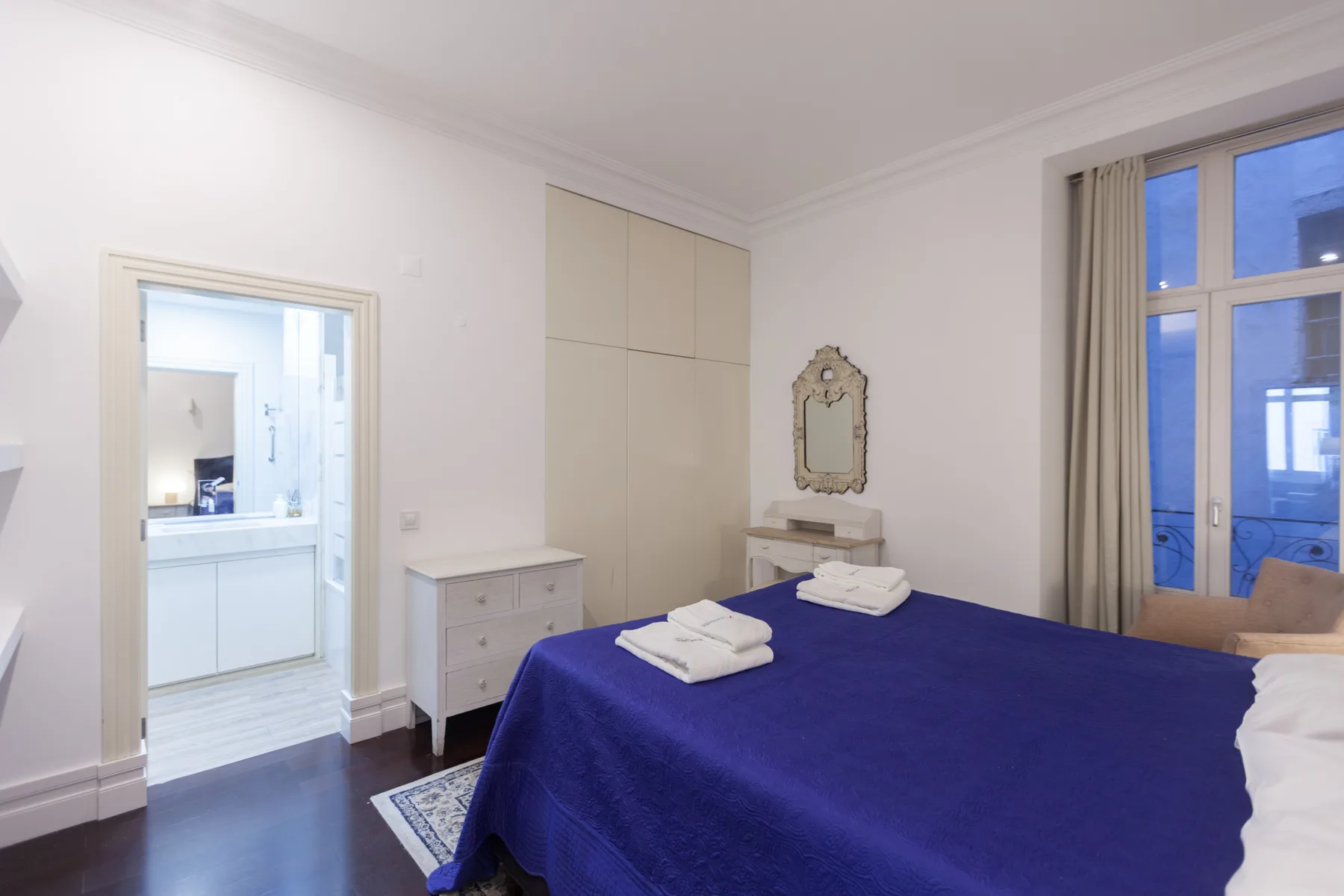 Un dormitorio con una cama cubierta de azul y una puerta abierta a un baño luminoso.