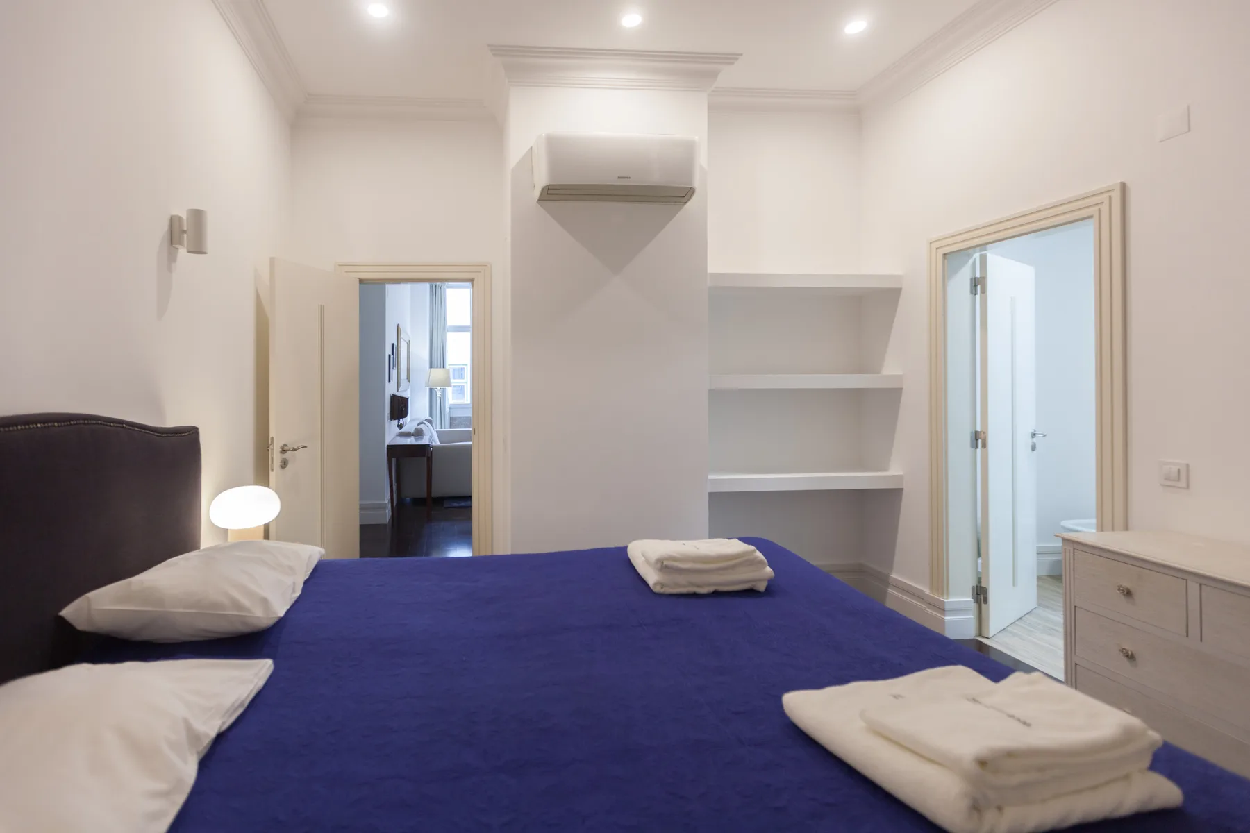Un dormitorio con una colcha azul, una lámpara de noche y estanterías empotradas.