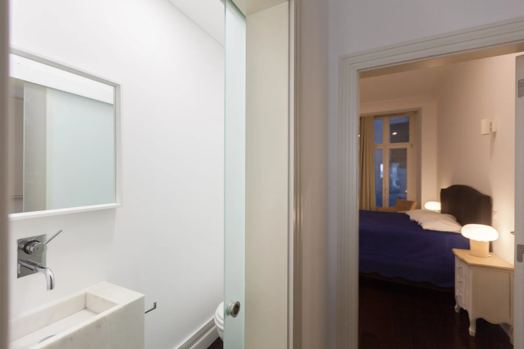 Un baño con lavabo y espejo da a un dormitorio con una cama.