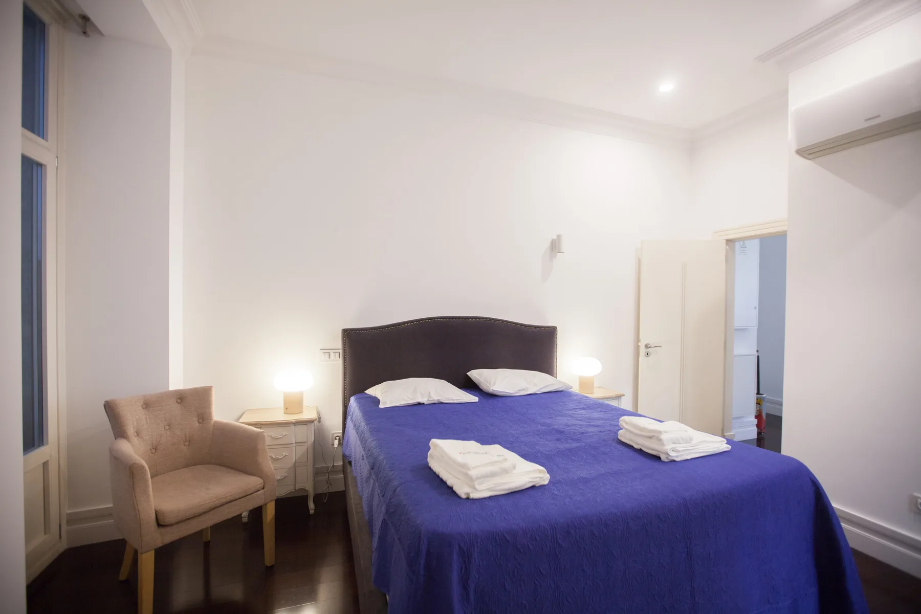 Un dormitorio con una cama cubierta de azul, mesillas y una silla tapizada.