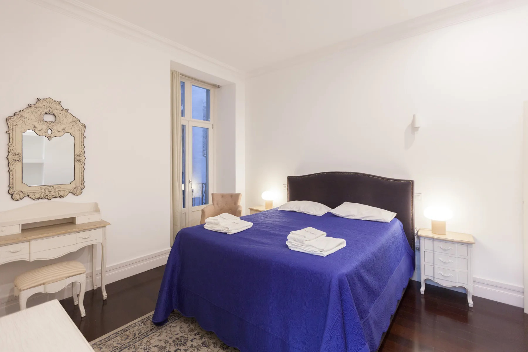 Un dormitorio con una cama cubierta de azul, mesitas de noche y un pequeño tocador.