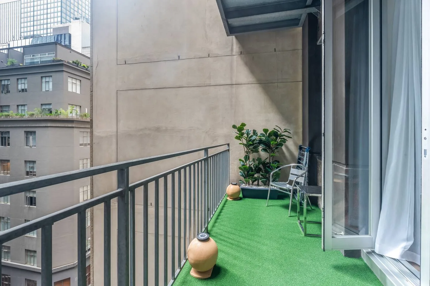 A balcony with green flooring, seating, and potted plants overlooks nearby buildings.