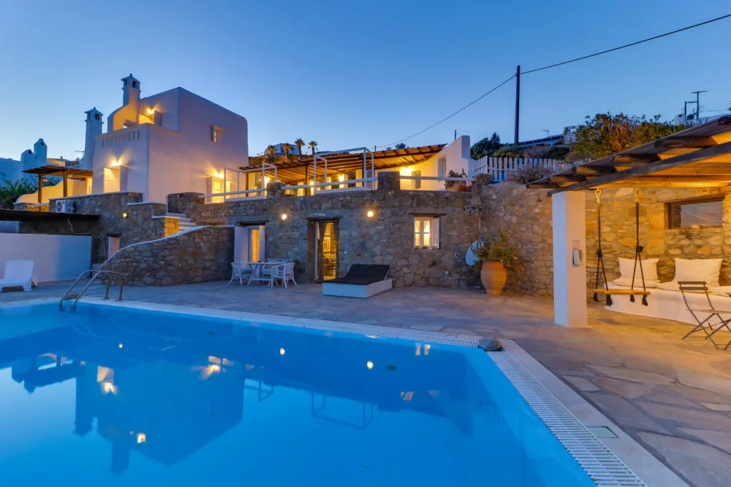 A swimming pool beside a stone terrace and illuminated villa at dusk.