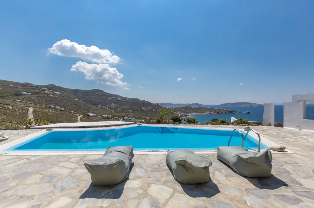 A swimming pool overlooks a coastal landscape, with cushioned seating on the surrounding terrace.