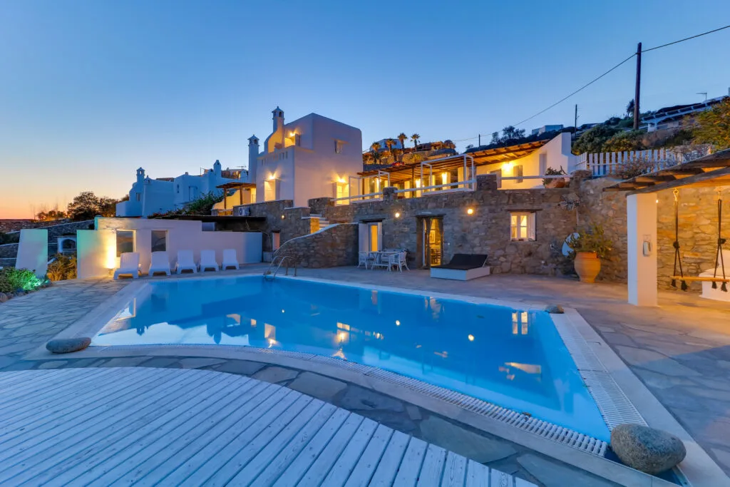 An outdoor swimming pool sits beside stone walls and illuminated buildings at dusk.