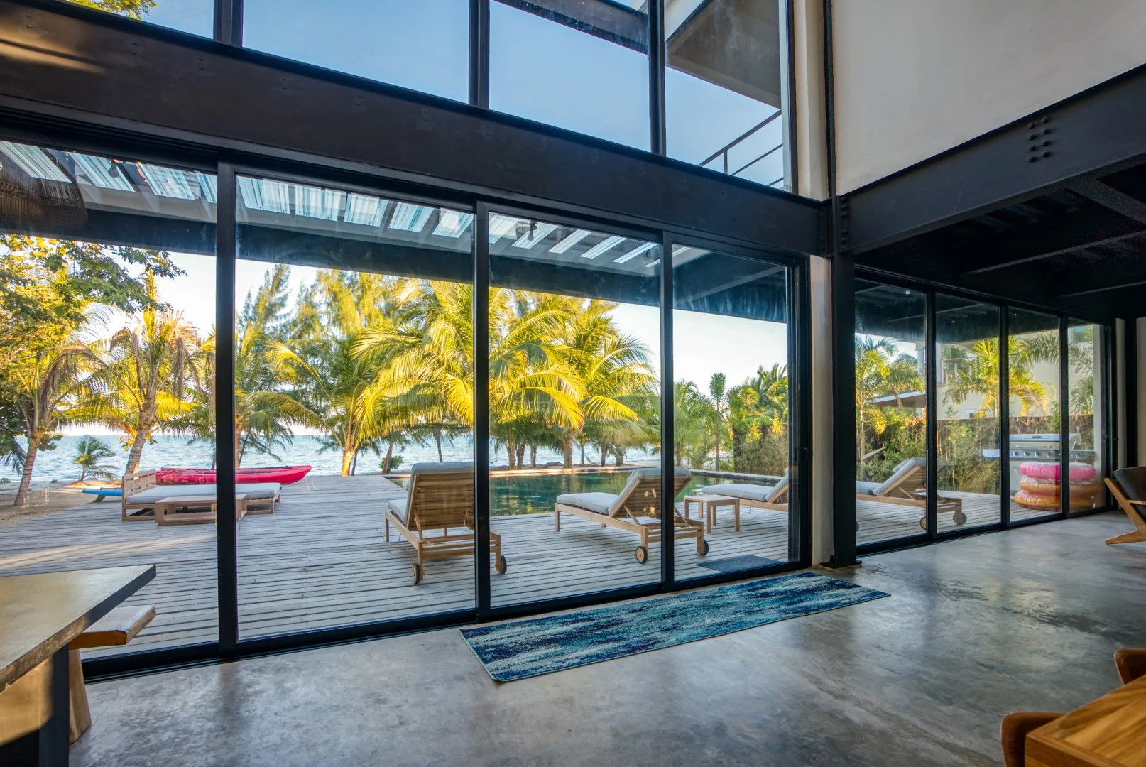 A glass-walled living area opens onto a wooden deck with palm trees beyond.