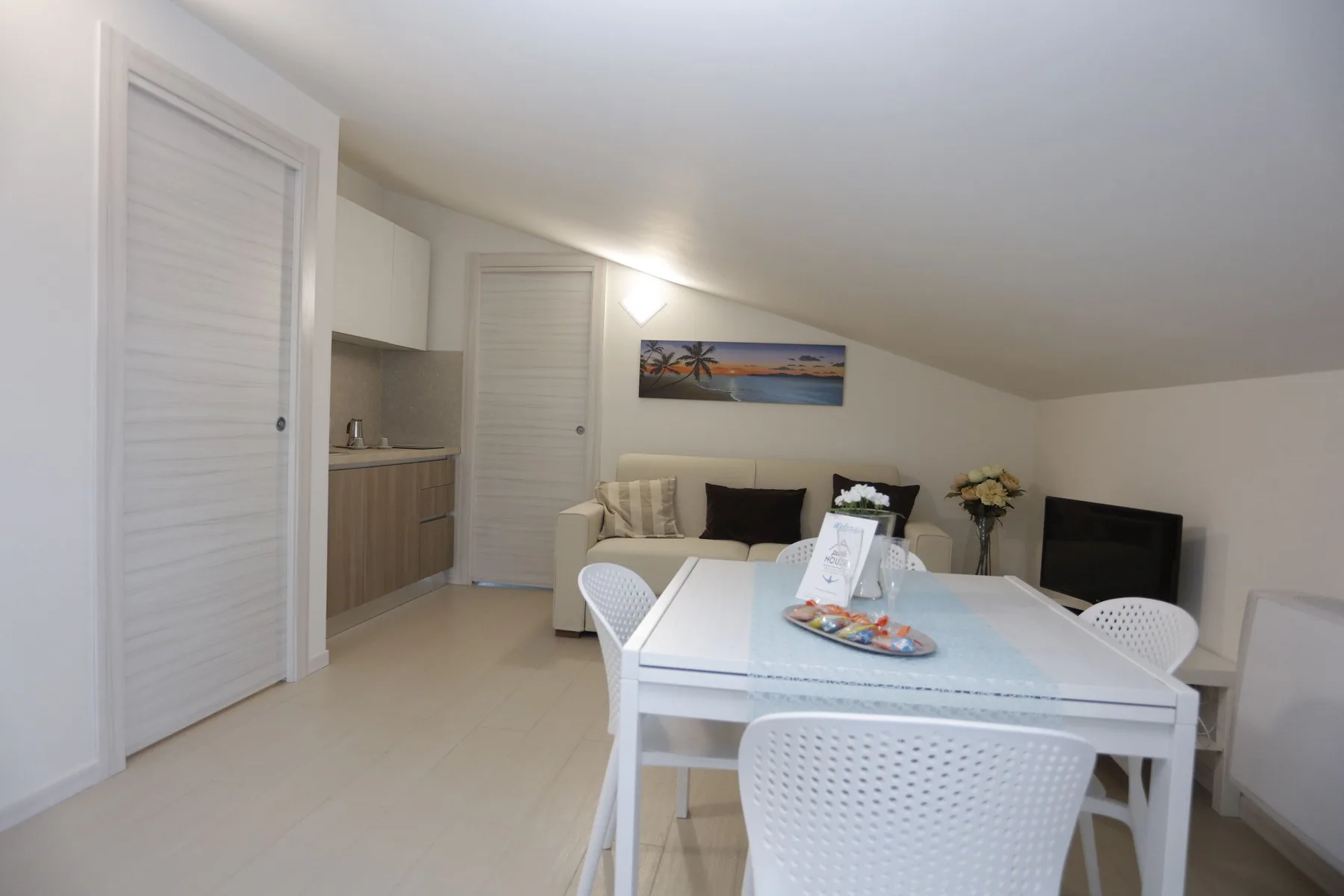 A dining area with a white table and chairs, beside a compact kitchenette and seating.