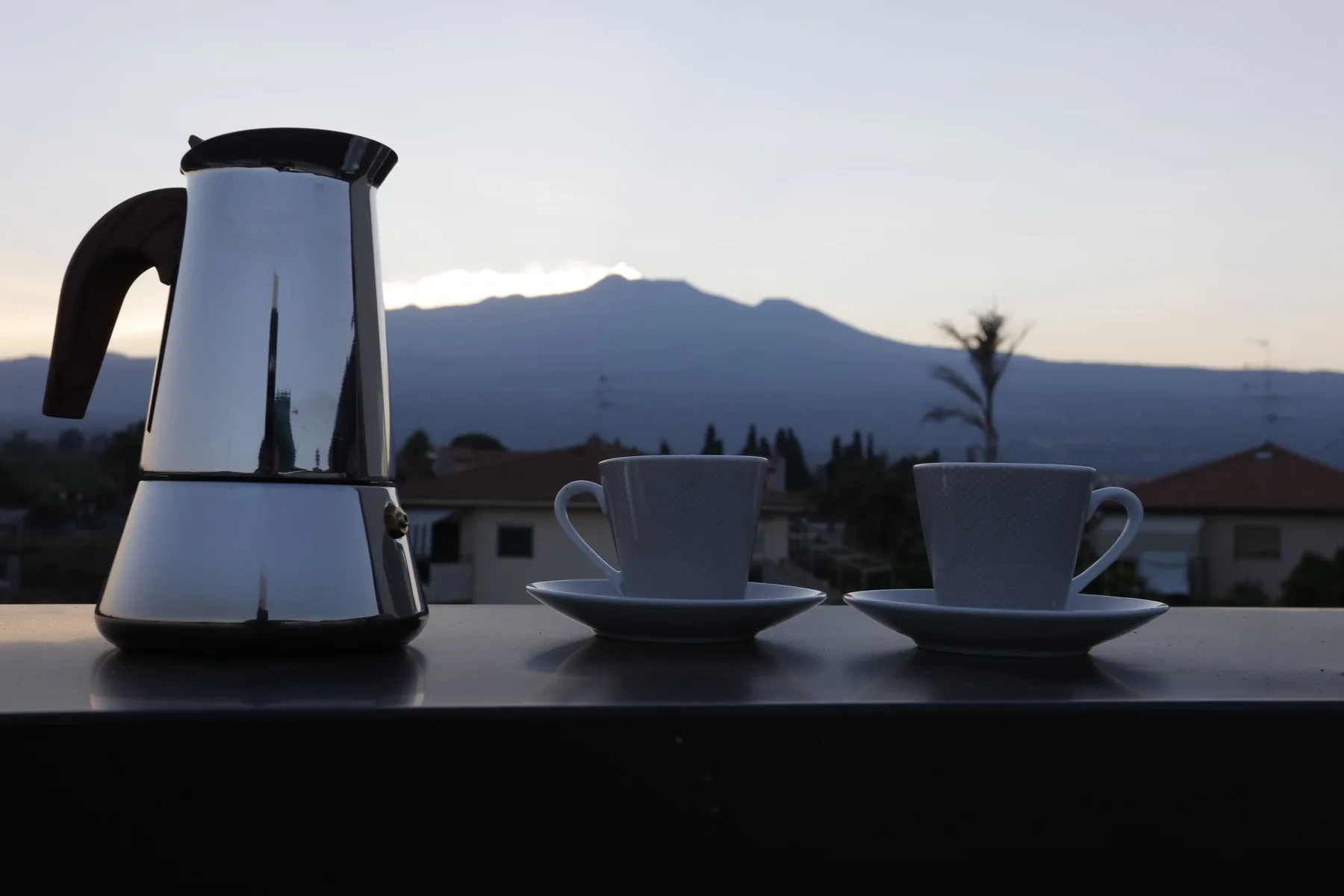 A coffee pot and two cups sit on a table with mountains visible in the distance.