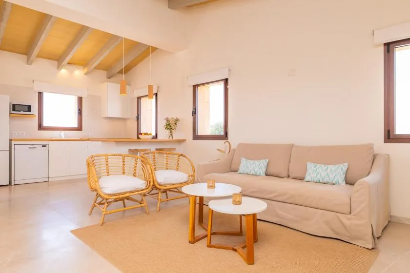 A bright living area with a beige sofa, wicker seating, and an open kitchen.