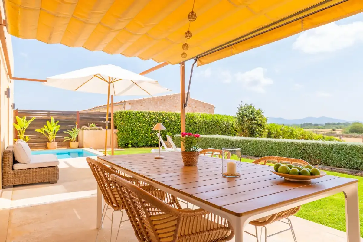 Outdoor dining area under an awning, with a long table and surrounding greenery.
