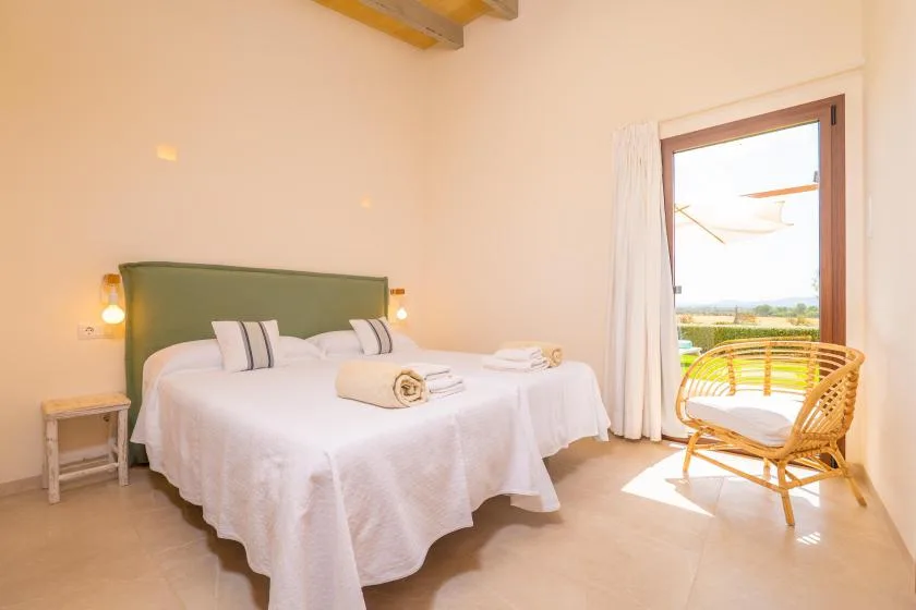 A bright bedroom with beds, white linens, and a wicker chair by the open doorway.