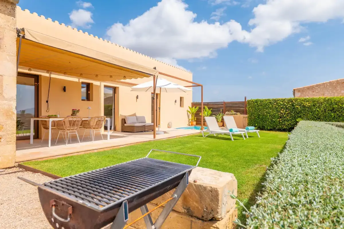 A sunny patio and lawn beside a house, with outdoor seating and a grill.