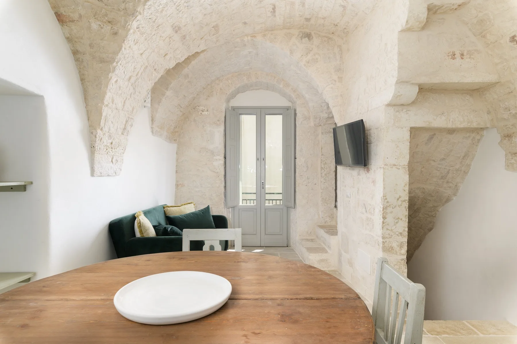 Un comedor con una mesa de madera, asientos y una ventana alta bajo arcos de piedra.