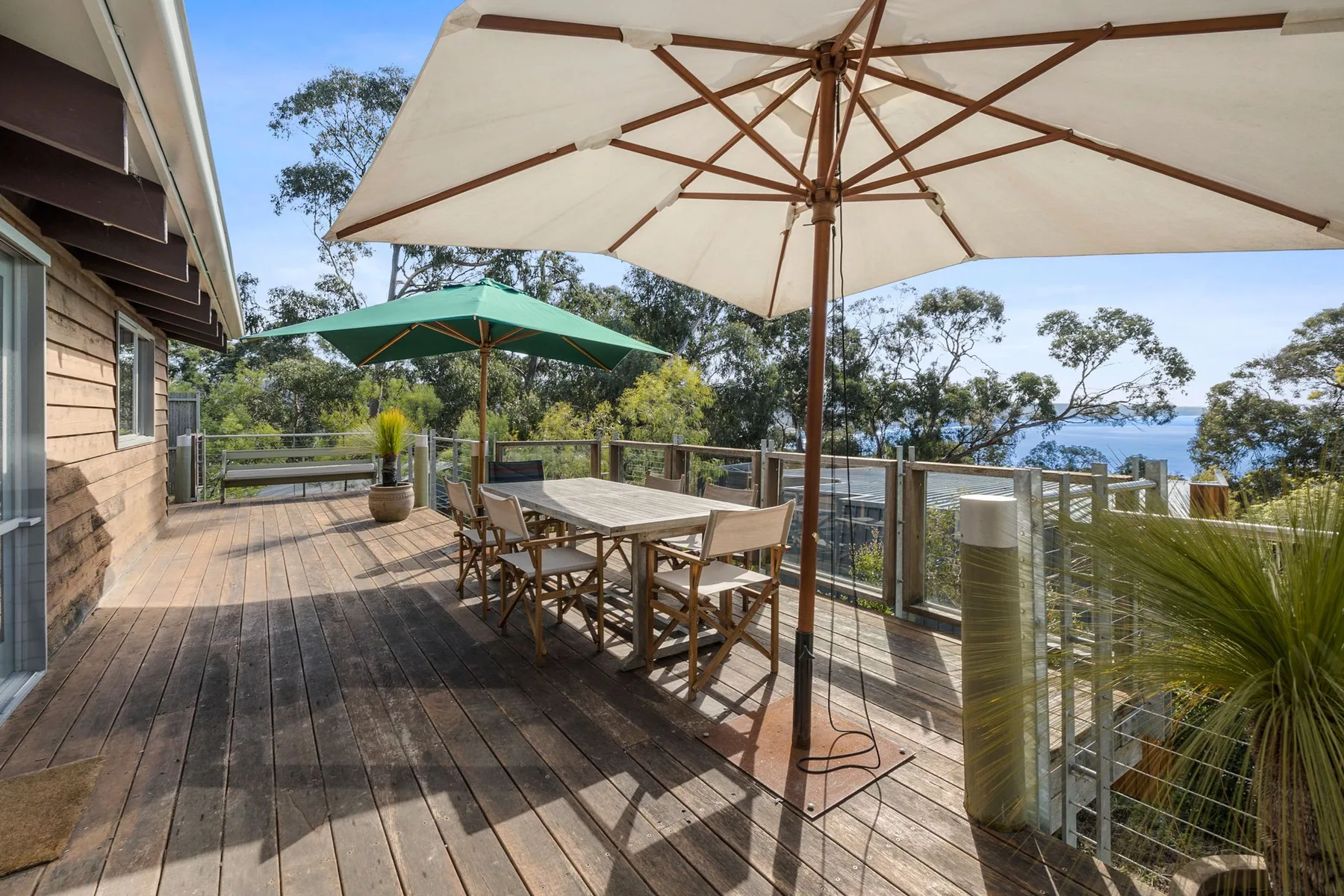An outdoor deck with a dining table and umbrellas overlooks trees and water.