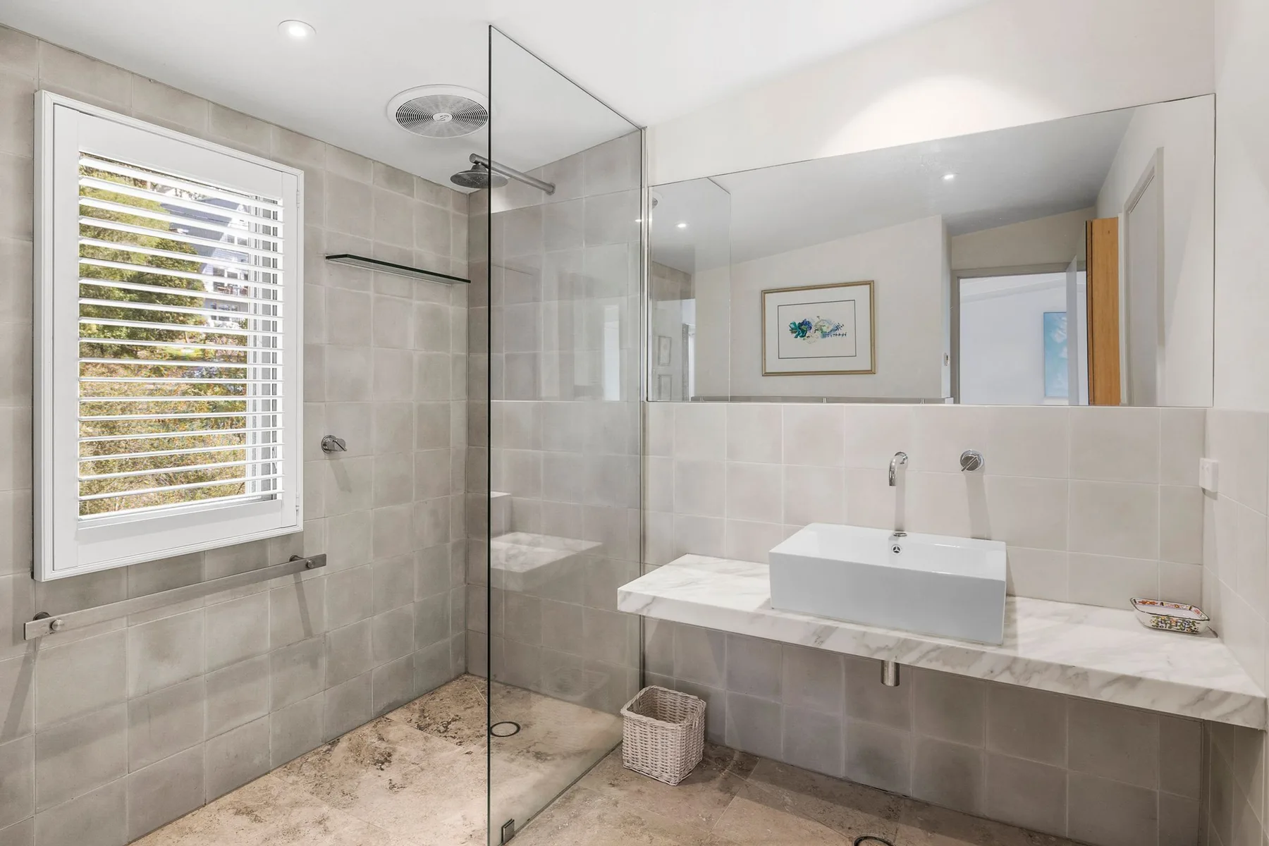 A tiled bathroom has a glass shower and a sink beneath a wide mirror.