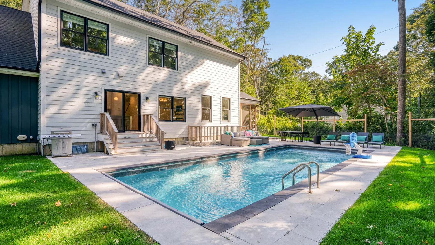 A swimming pool beside a light-colored house, with a patio and surrounding trees.