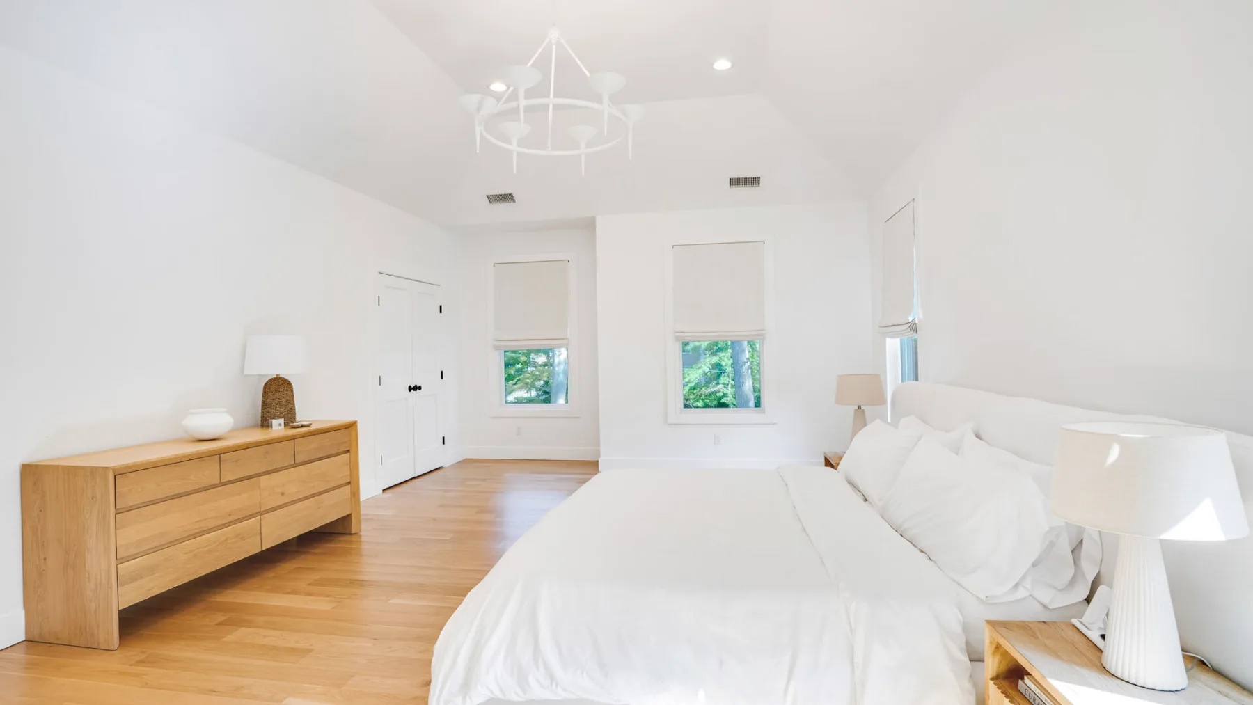A bright bedroom with a large bed, wooden dresser, and windows.
