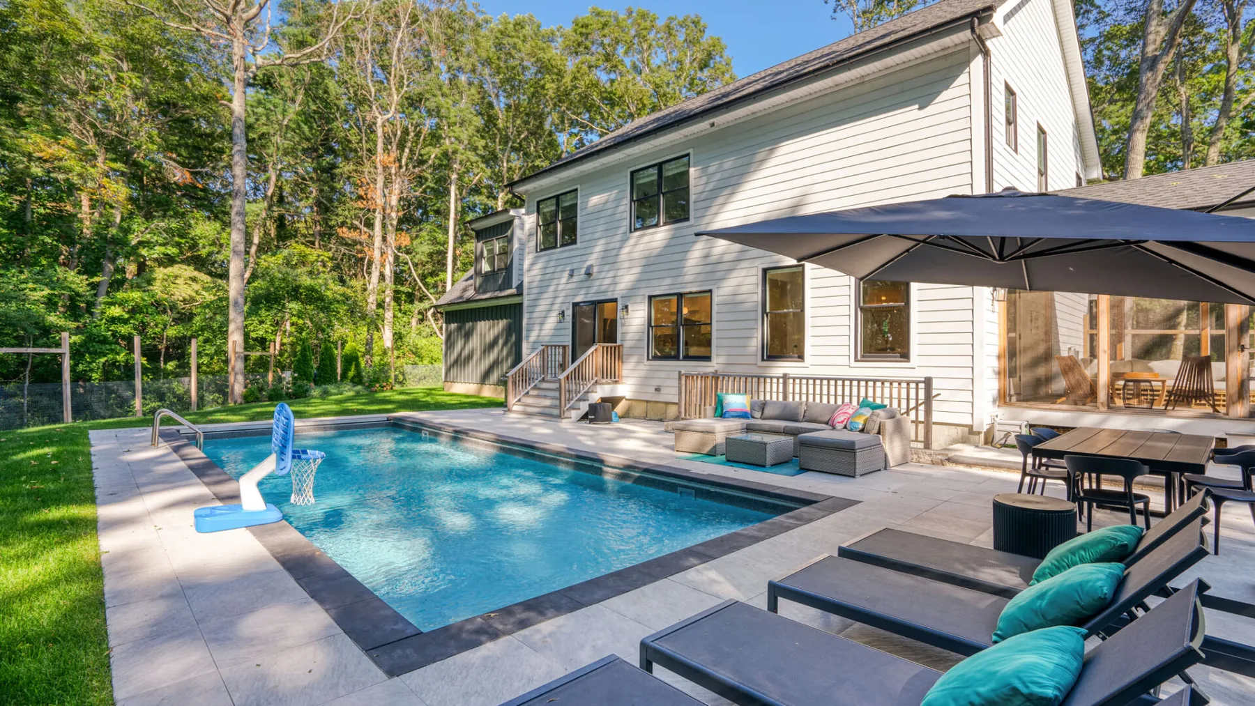 A backyard pool beside a white house, with outdoor seating and a patio umbrella.