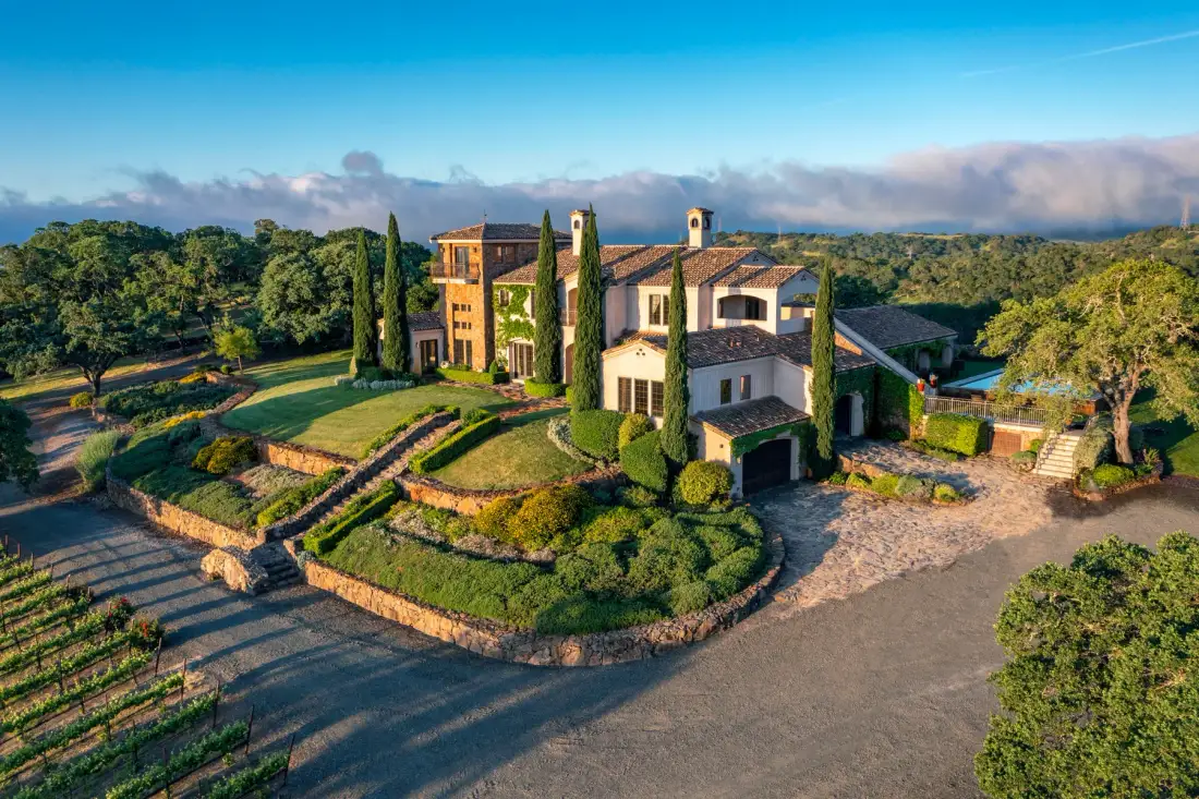A large villa sits among landscaped gardens and tall cypress trees.