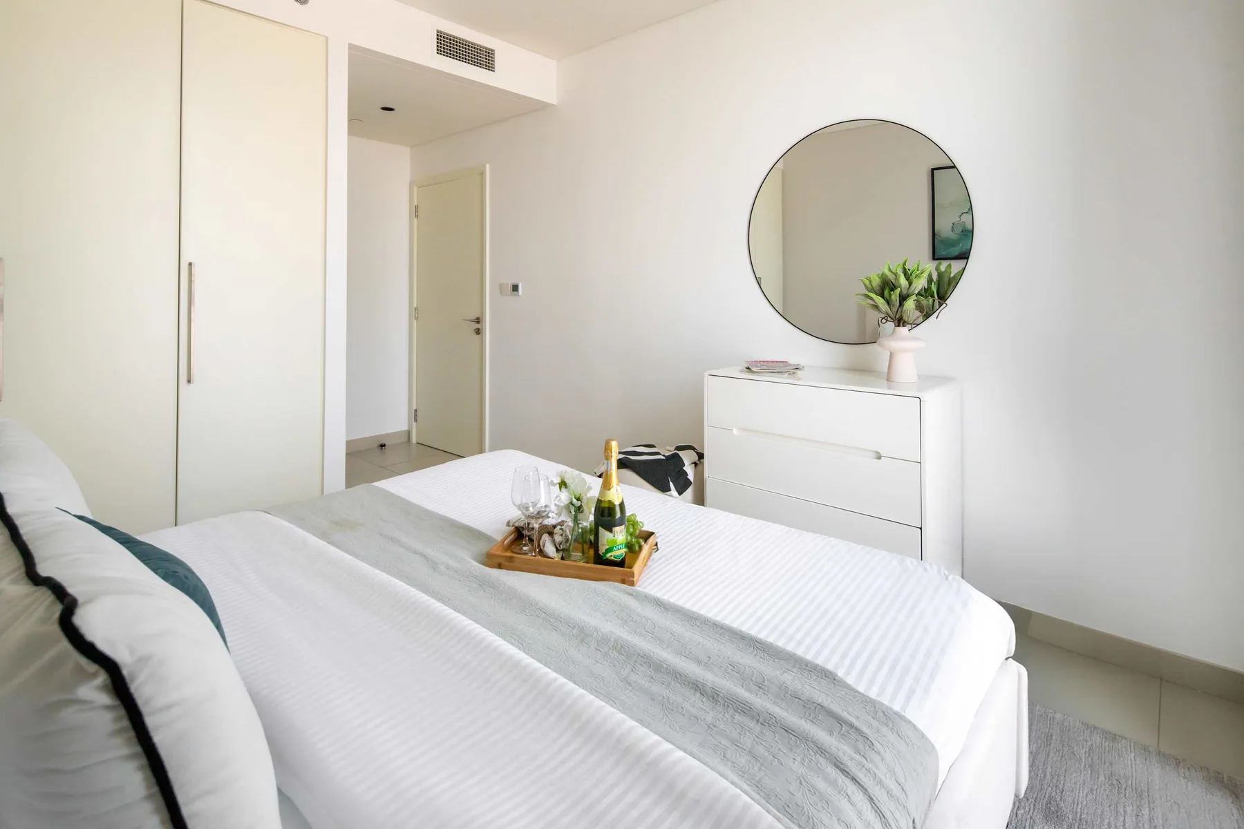 Un dormitorio luminoso con una cama bien hecha y una cómoda blanca con un espejo redondo.