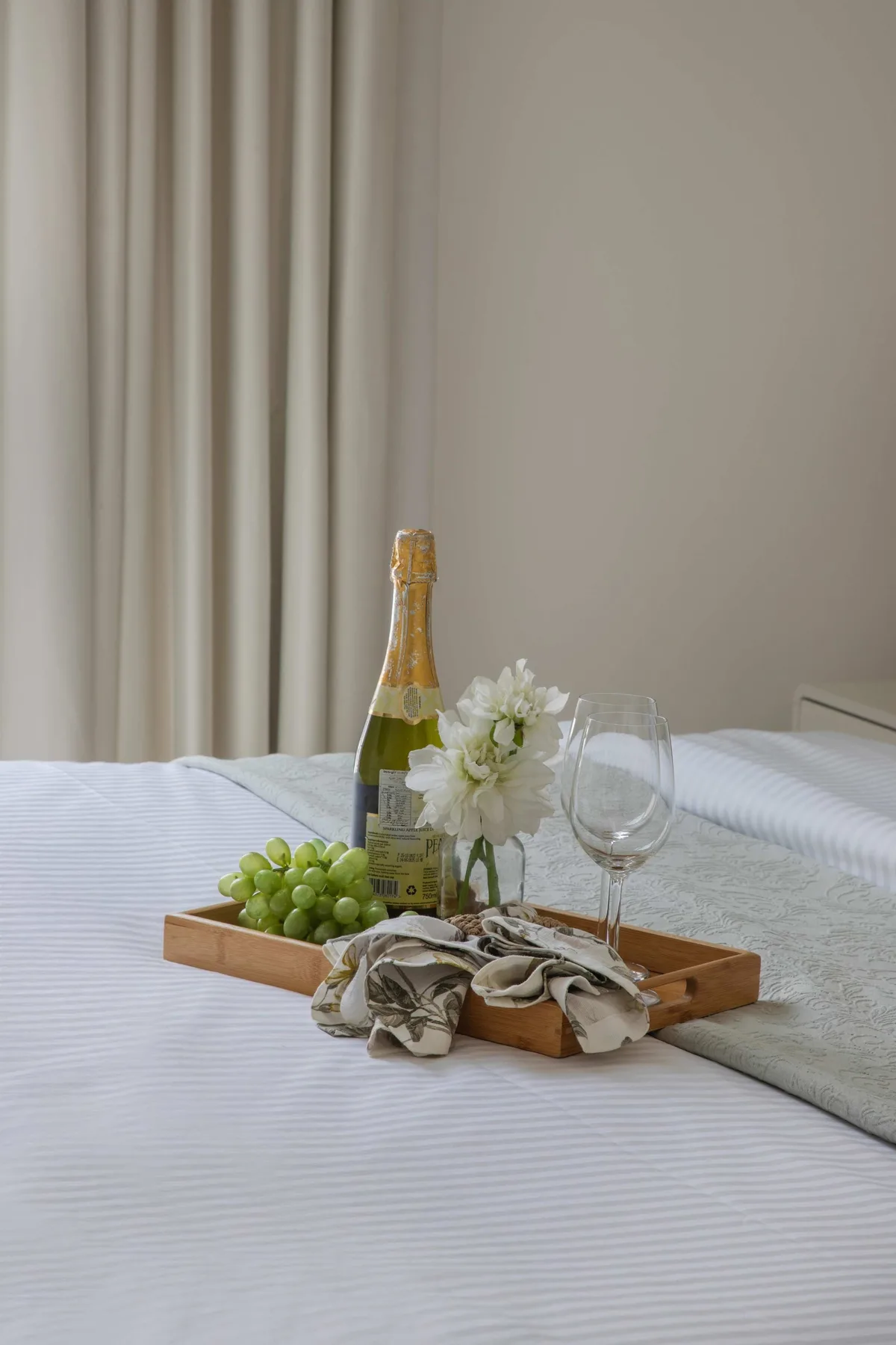 Una cama bien arreglada muestra una bandeja de madera con una botella, flores y copas.