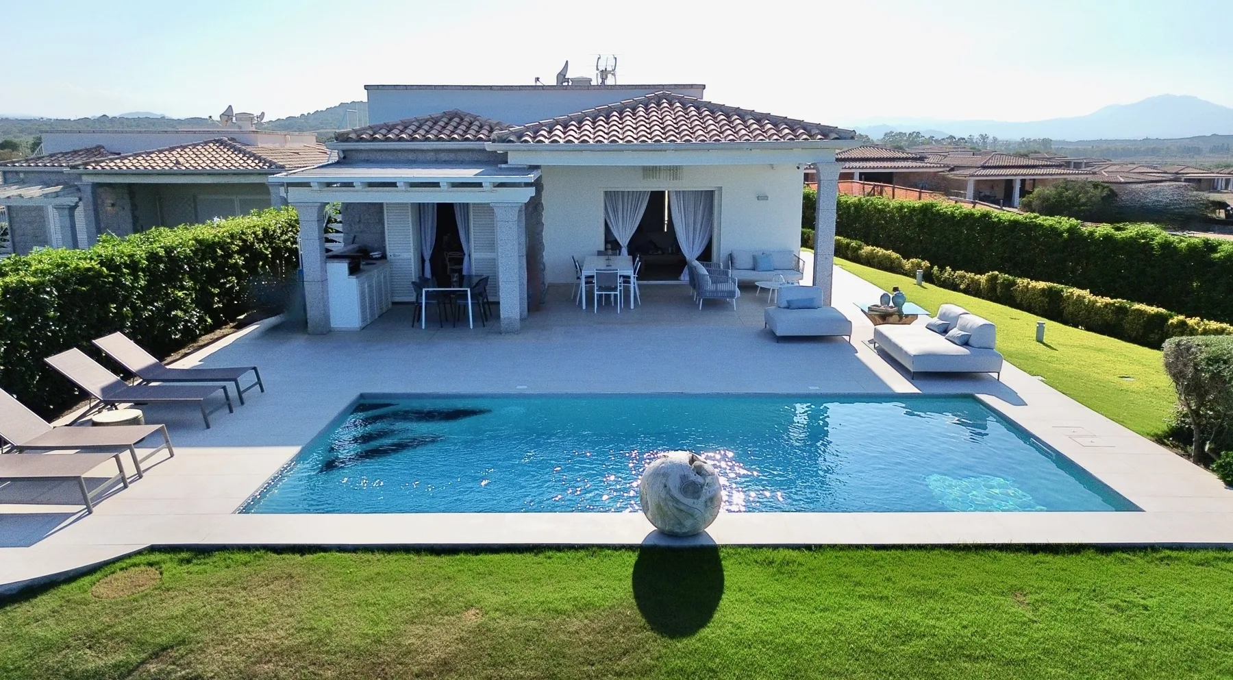 Una piscina y un patio están frente a una casa, con césped y asientos al aire libre.