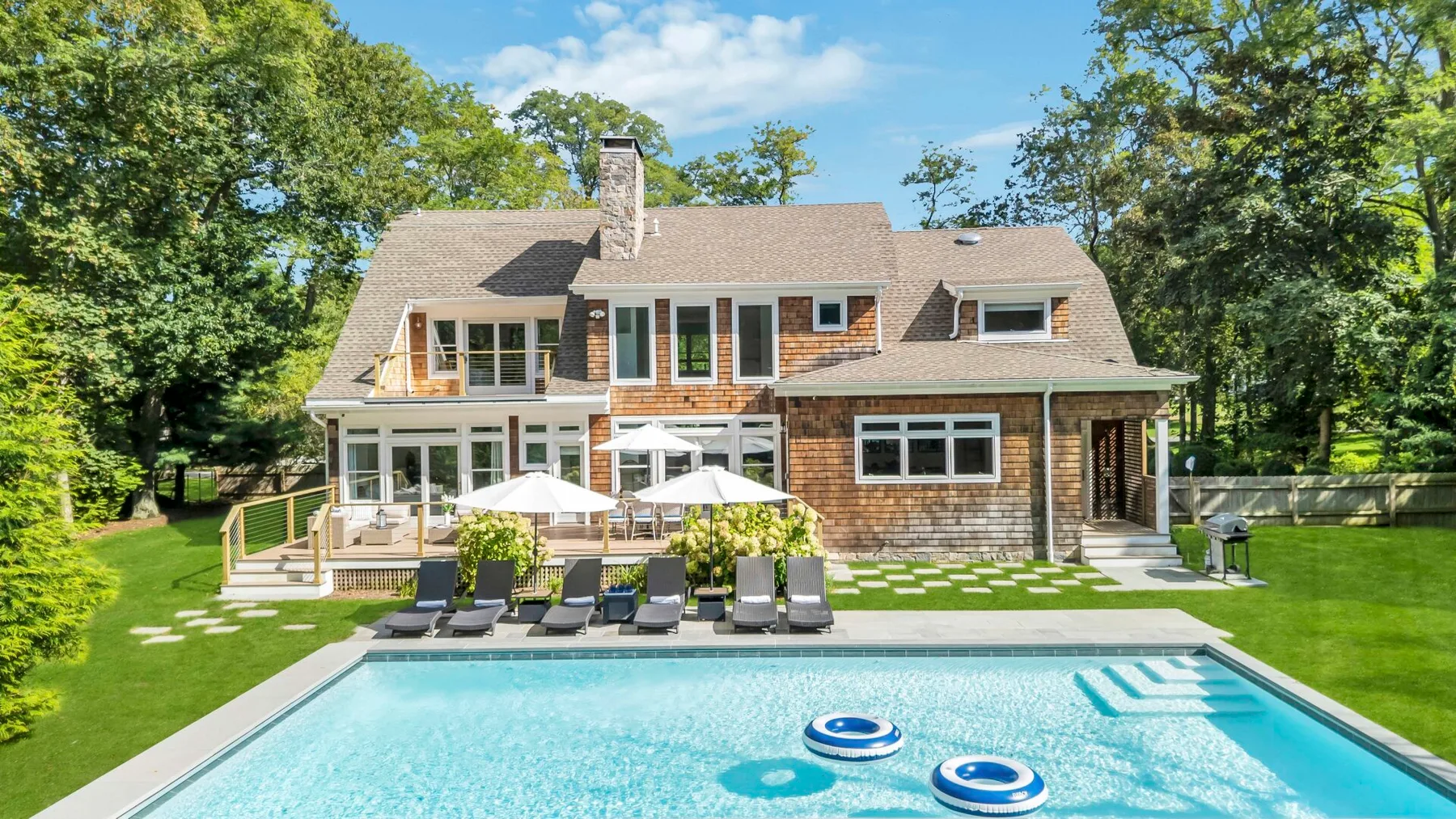 A large house overlooks a swimming pool, with patio seating and trees around the lawn.