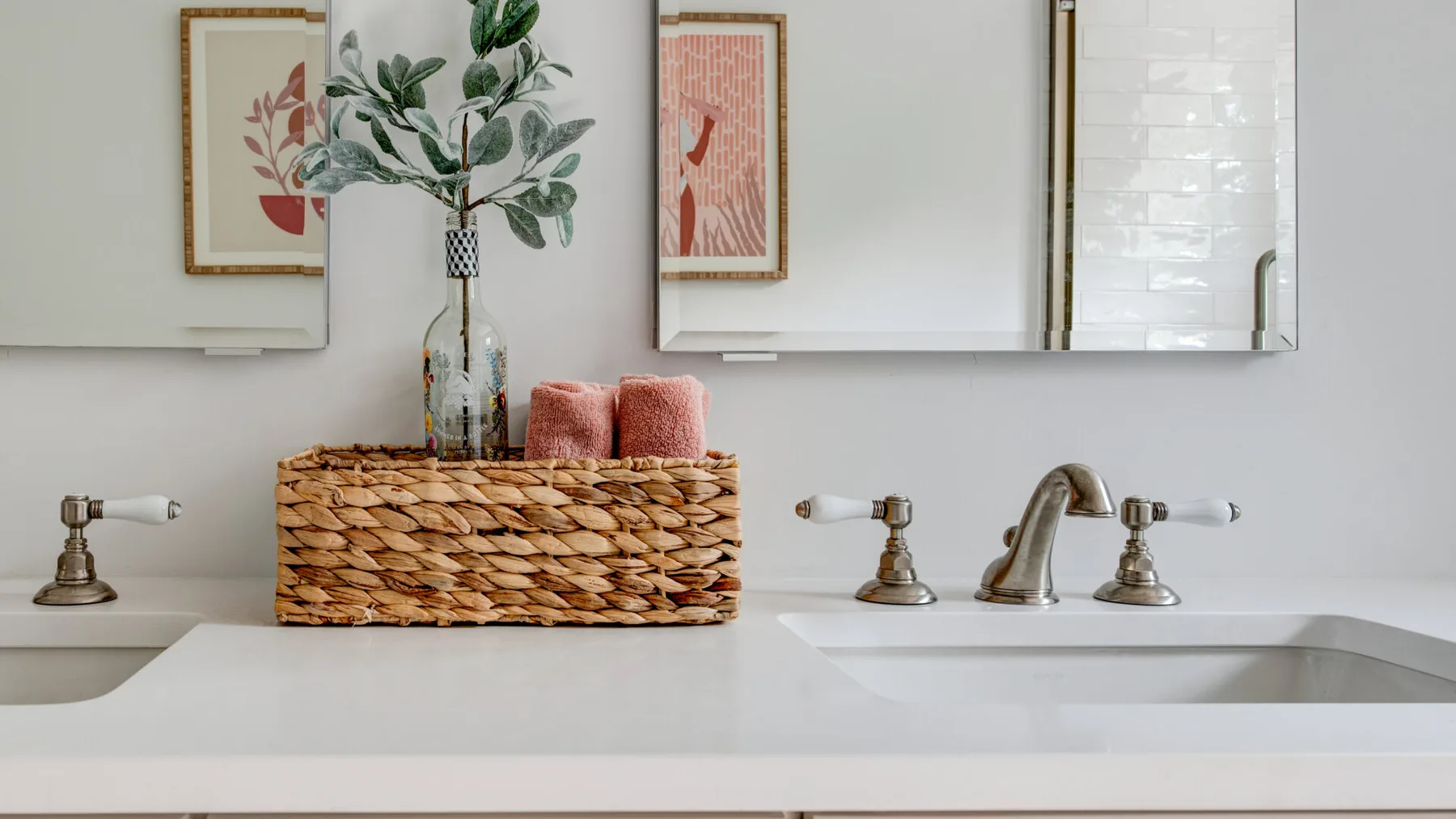 Un lavabo de baño con una cesta tejida, toallas y un jarrón con hojas bajo un espejo.