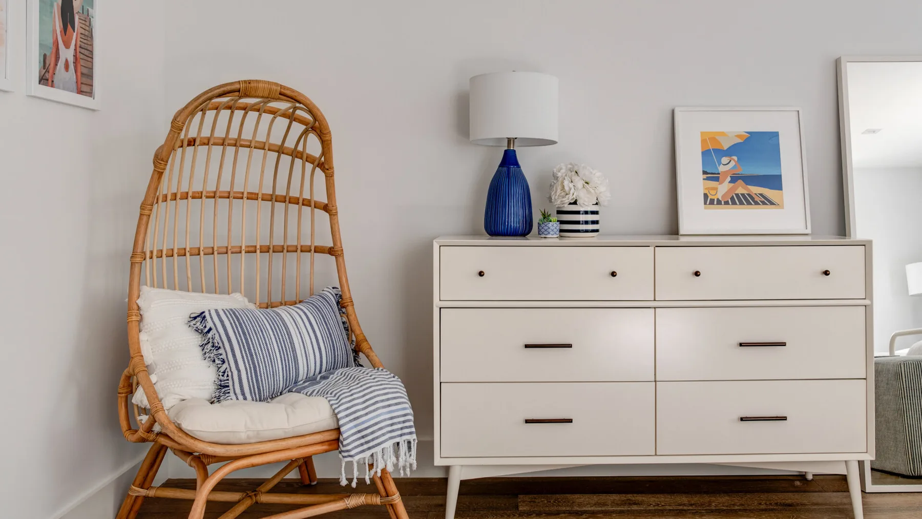 Un rincón luminoso de dormitorio con una silla de mimbre junto a una cómoda y cuadros enmarcados.