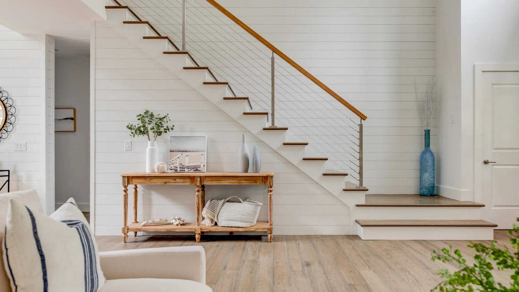 Una luminosa sala de estar con una escalera y una mesa consola junto a una pared blanca revestida.