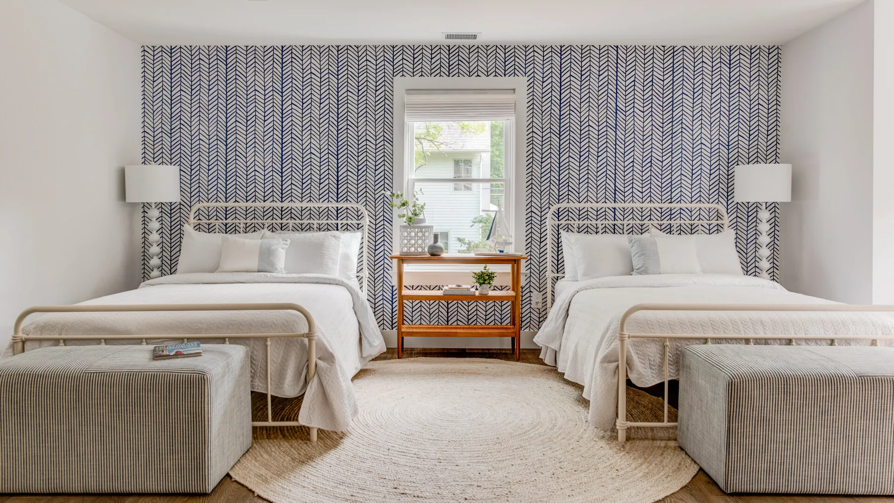 Un dormitorio con dos camas, papel tapiz estampado y una ventana sobre una mesa pequeña.