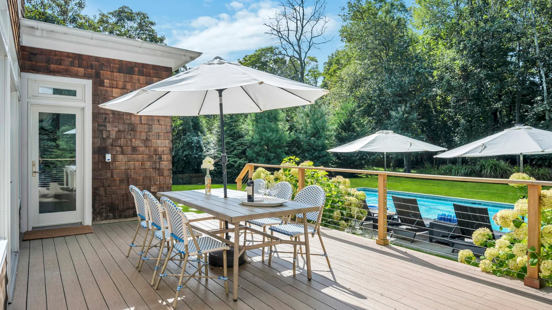 Comedor al aire libre en una terraza de madera junto a una piscina, con sombrillas.