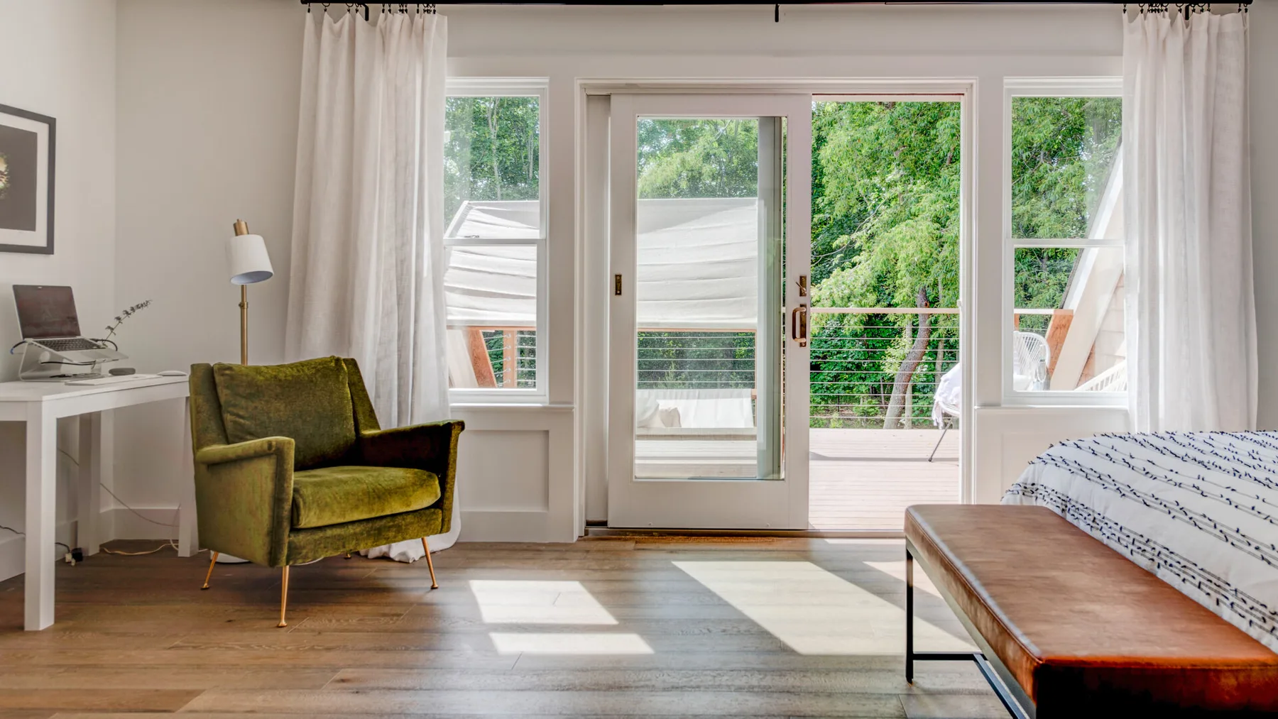 Un dormitorio luminoso con un sillón verde, una cama y puertas de cristal hacia la vegetación.