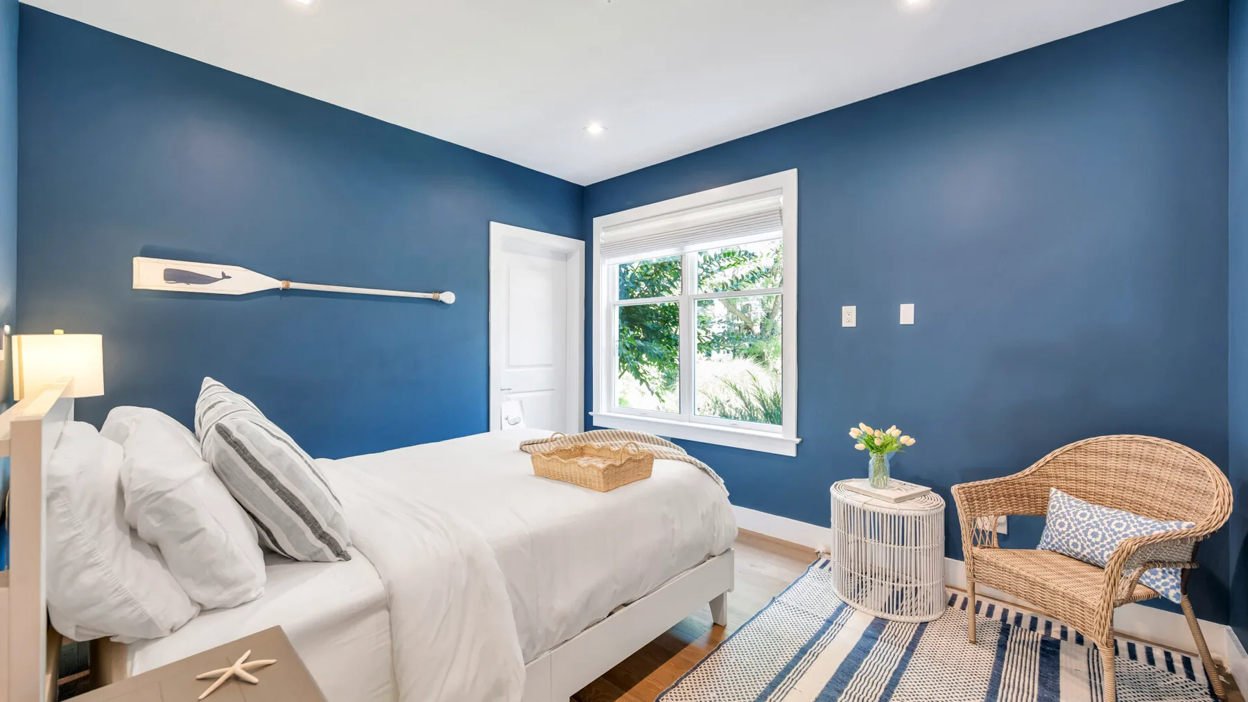 Un dormitorio con una cama blanca, paredes azules y una ventana con vistas a la vegetación.