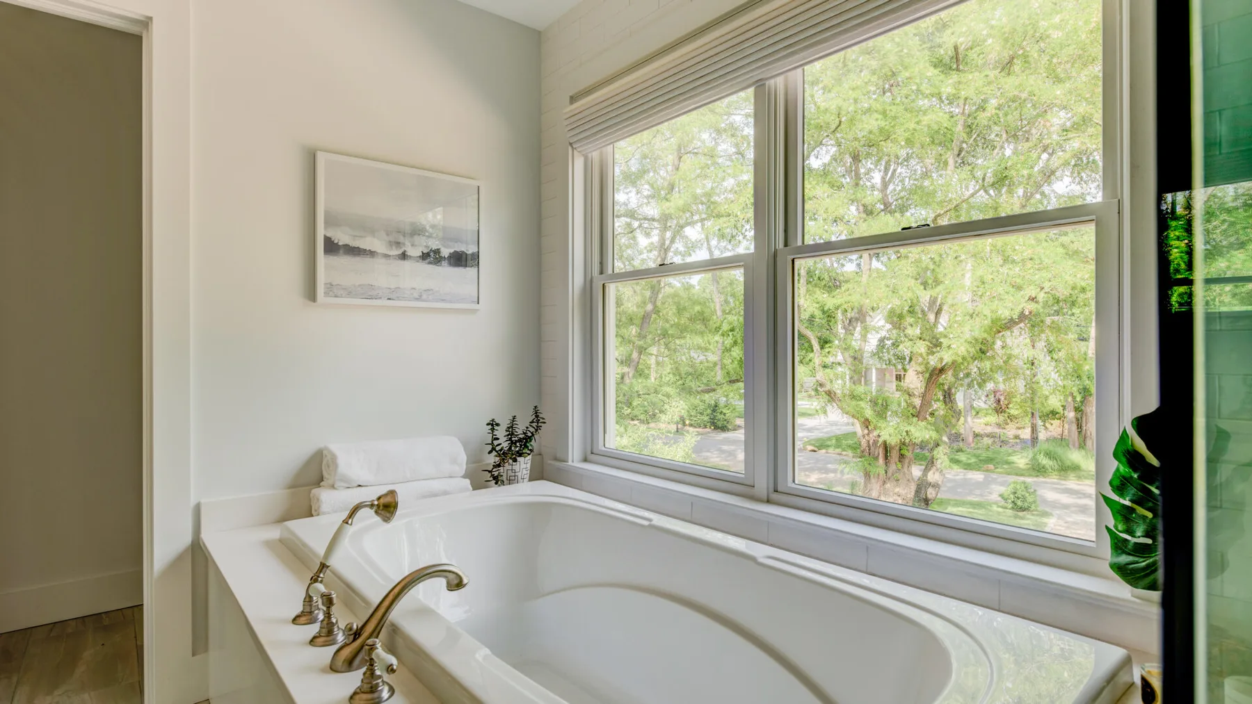 Un baño con bañera junto a grandes ventanas con vista a árboles frondosos.
